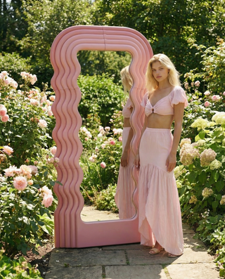 Woman in a pink dress standing next to a large pink mirror in a garden setting.