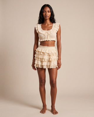 A woman stands barefoot against a plain warm beige studio background wearing a matching ruffled two-piece outfit with a cropped tank top and tiered shorts in a light cream or off-white color.
